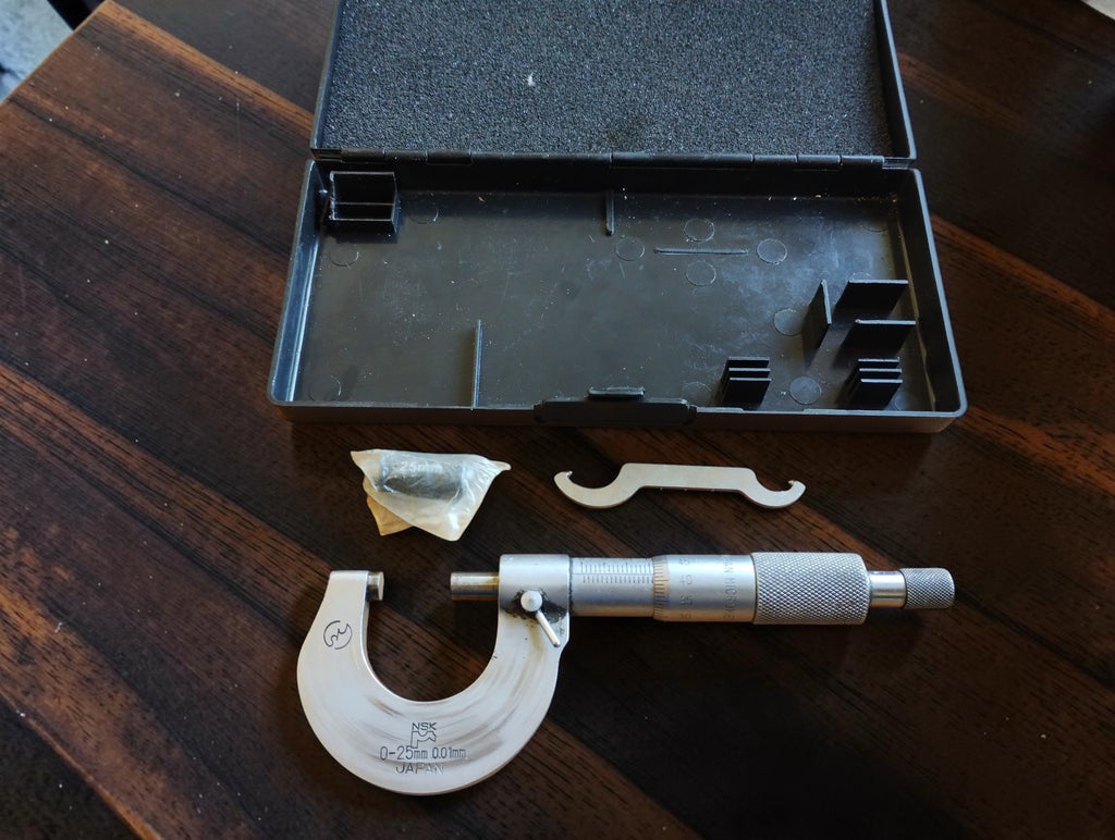Vintage NSK 0–25 mm outside micrometer with adjustment wrench, bagged accessory and open black storage case.

