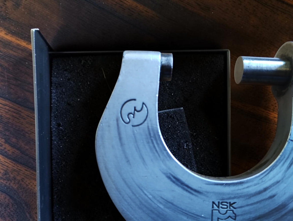 Close-up of the NSK micrometer's anvil, spindle and scratched metal frame with an engraved symbol.
