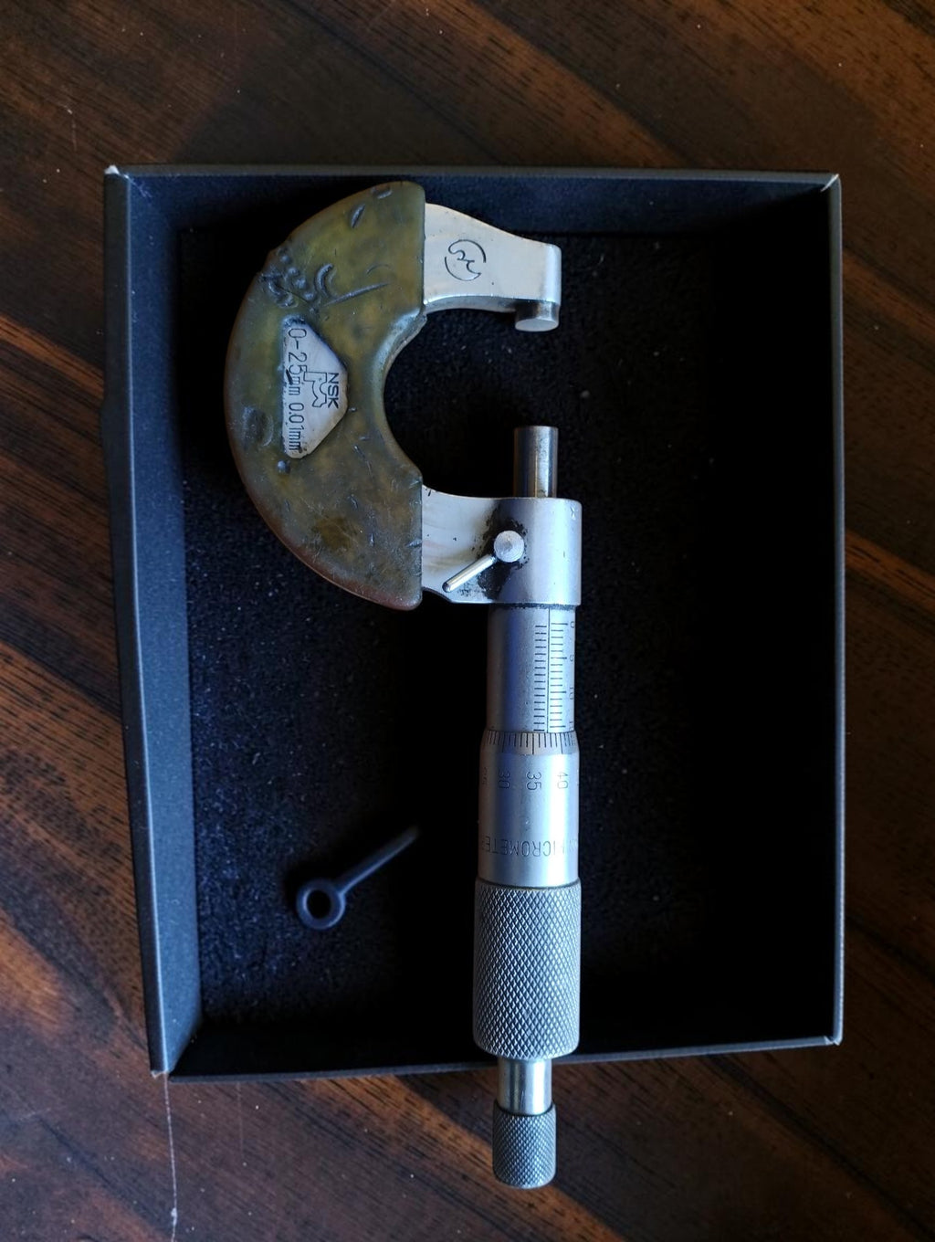 Vintage NSK 0–25 mm micrometer with discolored frame cover, graduated scale and knurled thimble in a black tray.
