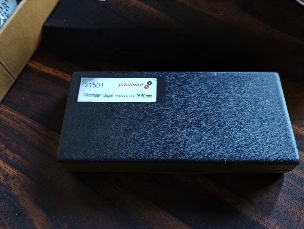 Closed black plastic storage case with a Paulimot 25–50 mm label that differs from the enclosed NSK 0–25 mm micrometer.
