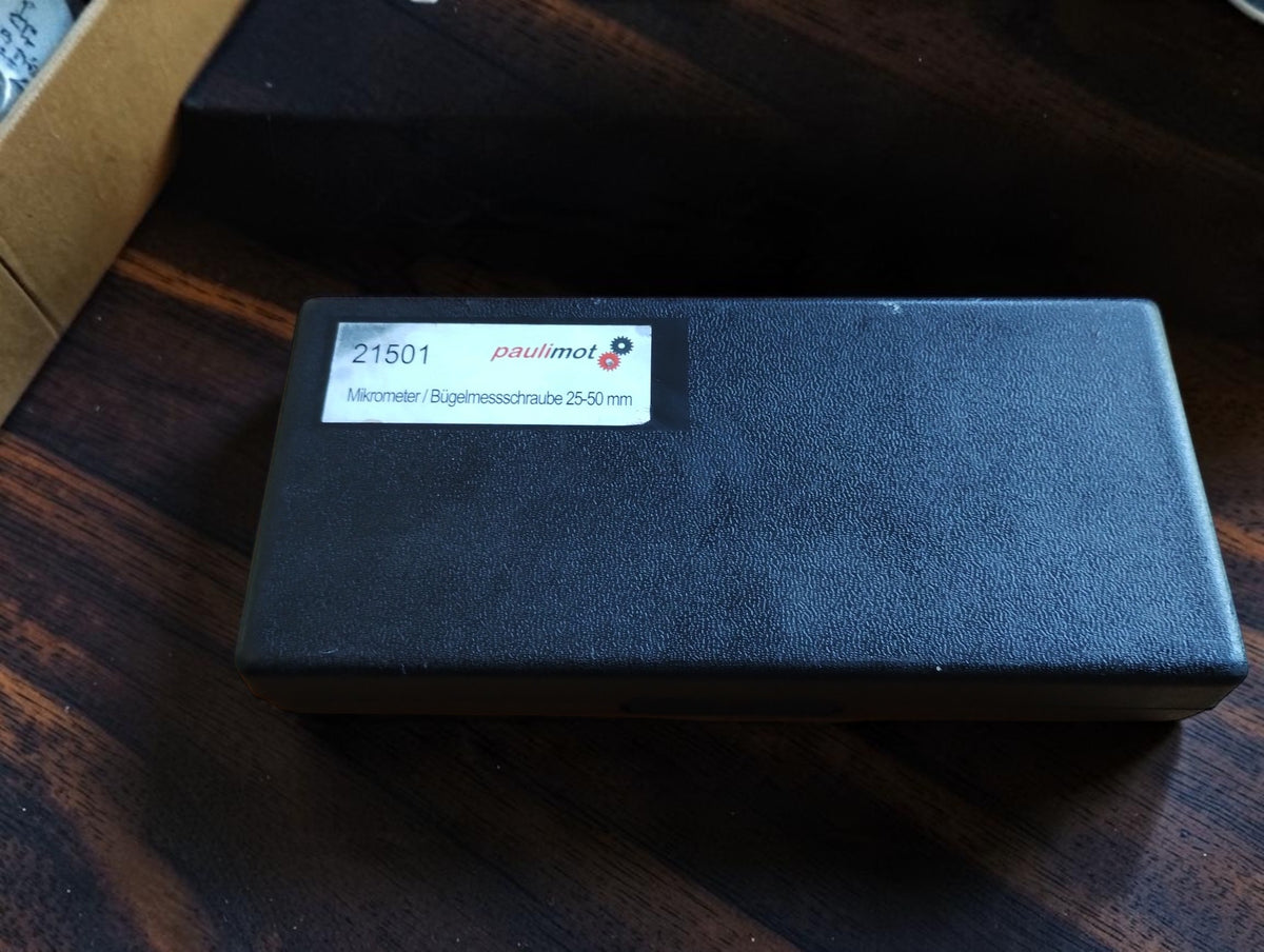 Closed black plastic storage case with a Paulimot 25–50 mm label that differs from the enclosed NSK 0–25 mm micrometer.
