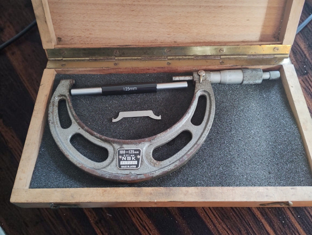 Vintage NSK 100–125 mm outside micrometer in an open wooden case with a 125 mm reference rod and adjustment wrench.
