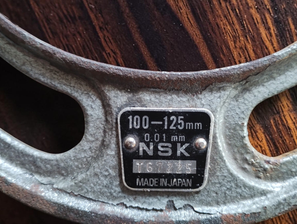 Close-up of the NSK micrometer nameplate marked 100–125 mm, 0.01 mm and Made in Japan, with worn paint on the frame.

