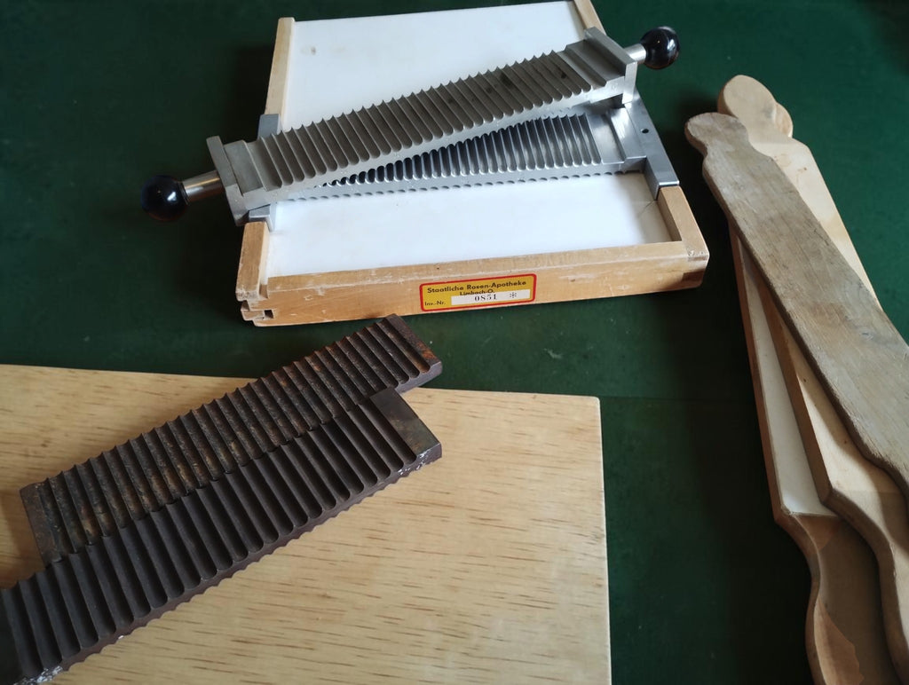 Angled view of the labeled Rosen-Apotheke base with grooved metal components, loose dark plates and worn wooden tools.
