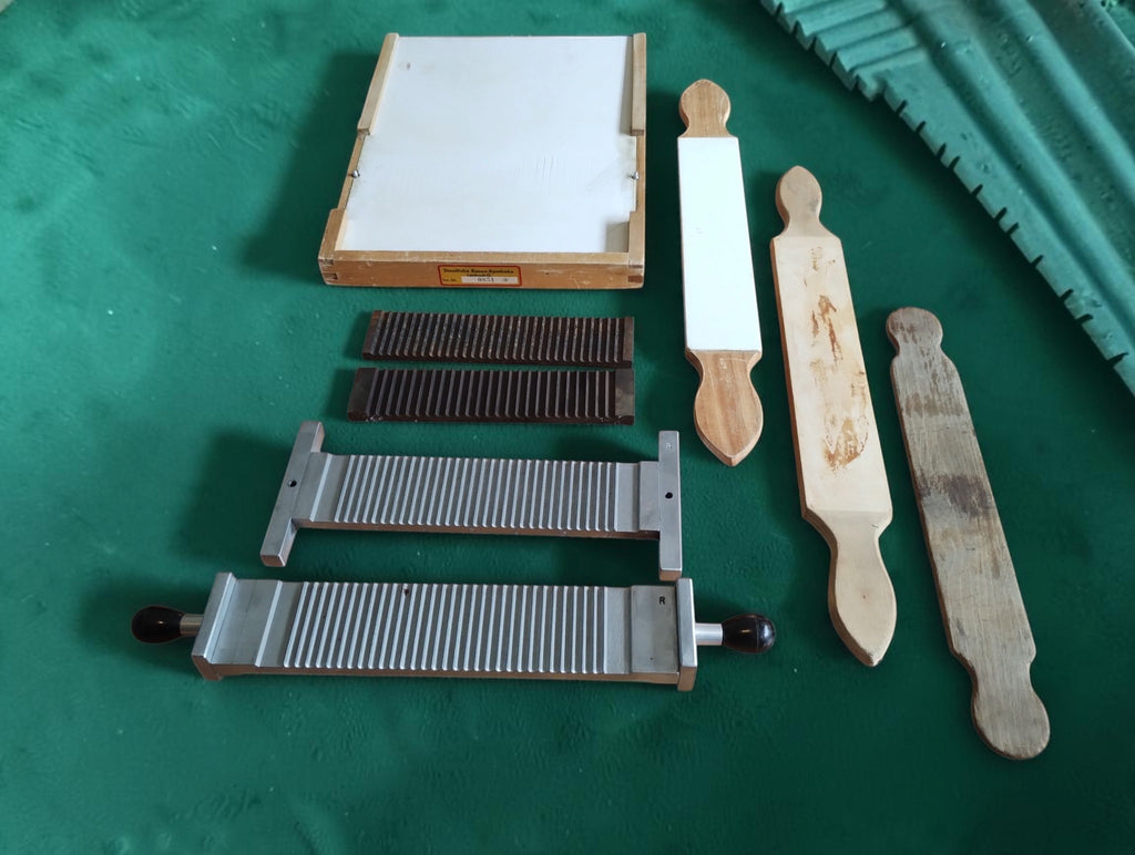 Wooden base with white inset, two dark grooved plates, silver-colored pill-making components and three wooden tools laid out separately.
