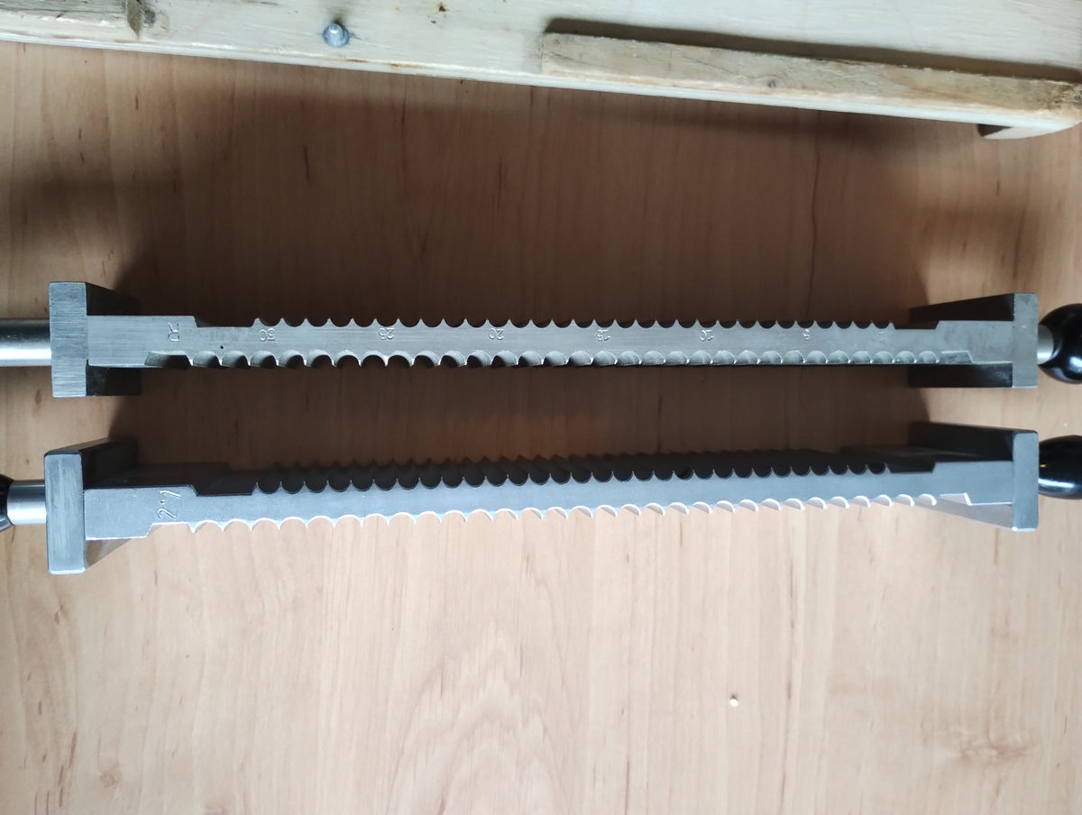 Side view of two grooved metal pill-making components with dark handles and engraved markings, resting on a wooden base.
