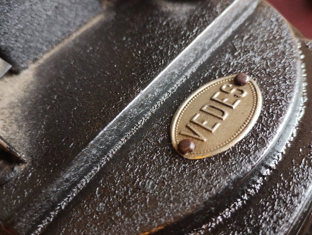Close-up of the riveted VEDES nameplate on the textured black base of the antique hand-cranked projector.
