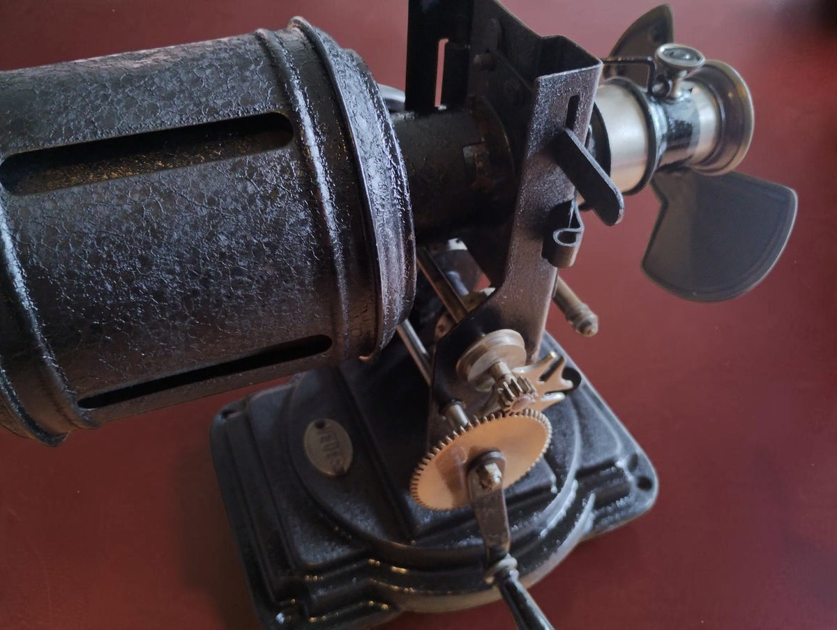 Angled view of the antique VEDES projector showing the slotted cylindrical housing, gears and hand crank.
