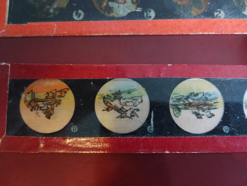 Close-up of a red-framed magic lantern glass slide with circular illustrated scenes and printed numbers.
