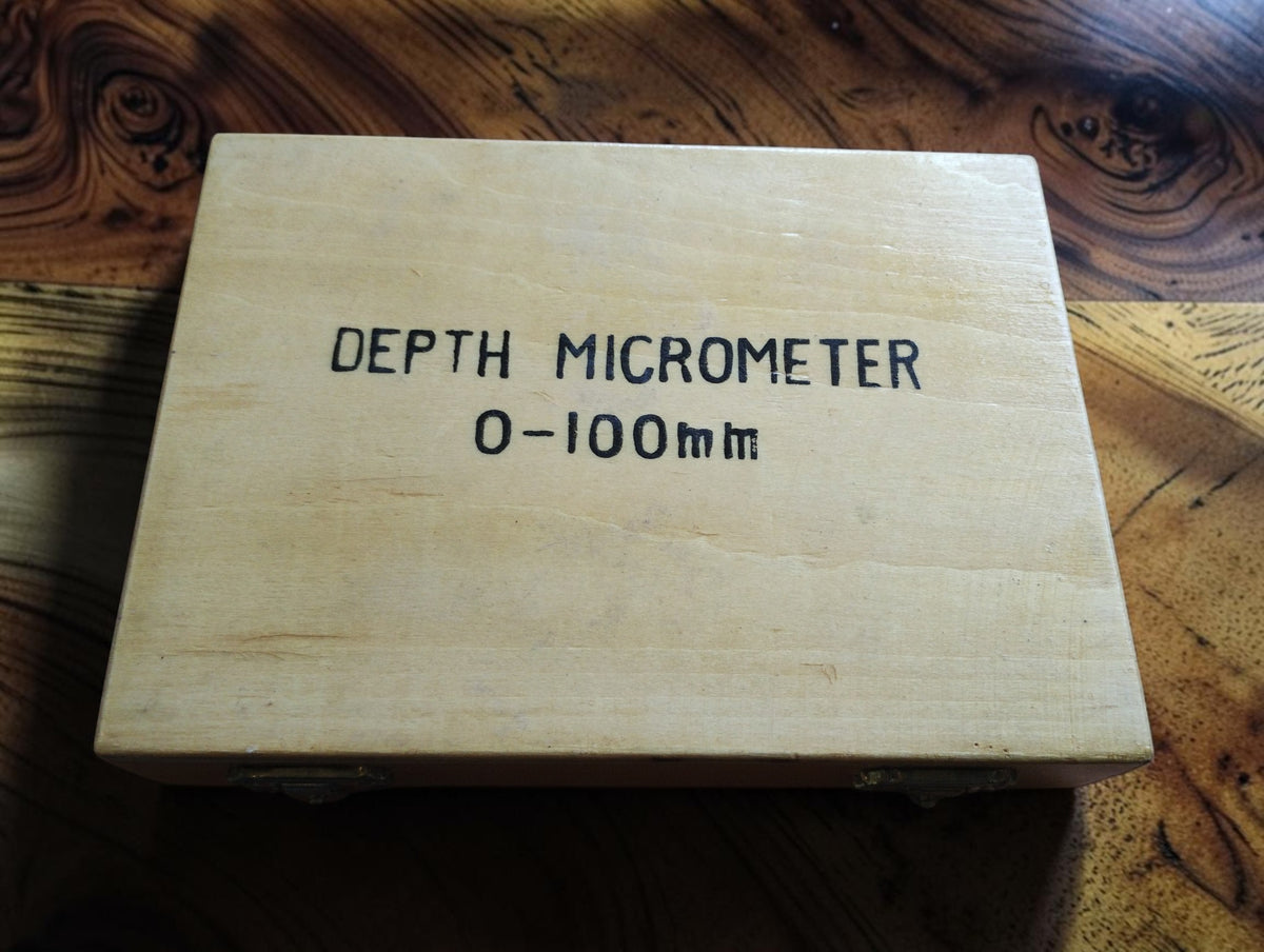 Analog depth micrometer standing on its flat metal base, shown outside the wooden case.
