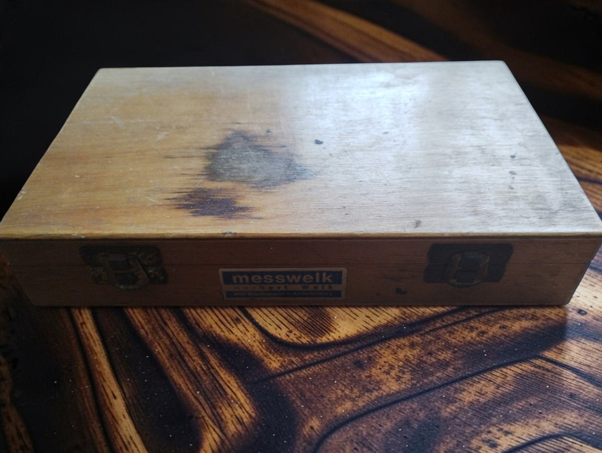 Closed wooden Messwelk micrometer case with metal clasps, front label and visible stains on the lid.
