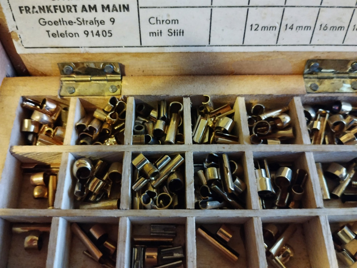 Close-up of brass-colored and silver-colored Riemenstifte fittings in the left compartments of the wooden box.
