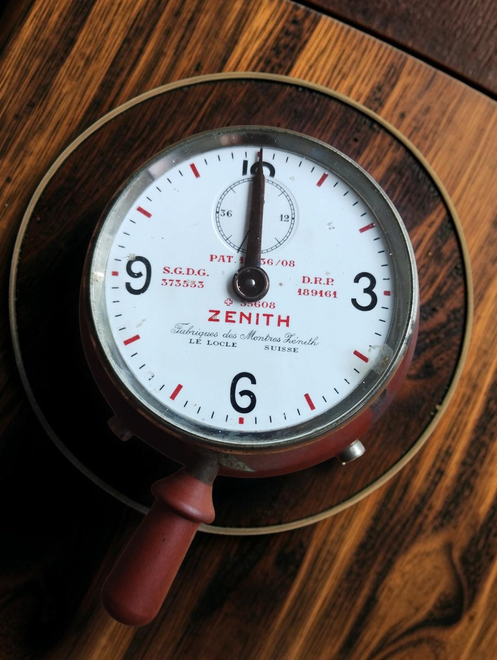 Antique Zenith Le Locle telephone timer with white dial, red markings, metal bezel and reddish-brown operating handle, angled view.
