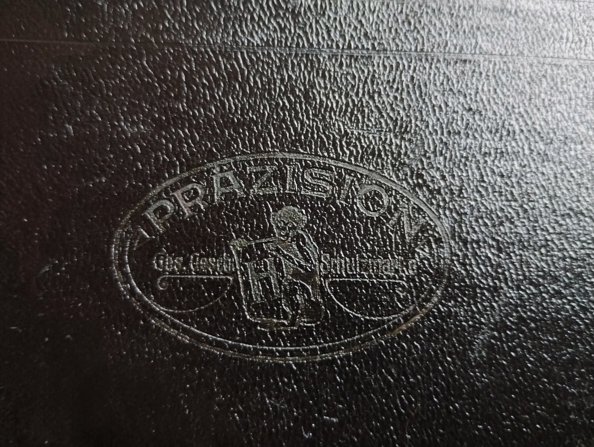 Close-up of the embossed oval PRÄZISION emblem with a central figure and shield on the black drafting instrument case.
