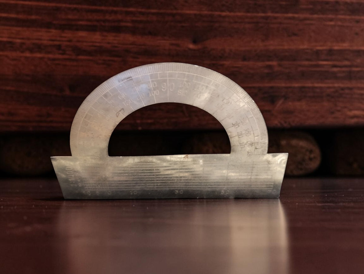 Metal semicircular protractor with engraved graduations, shown separately from the drafting instrument case.
