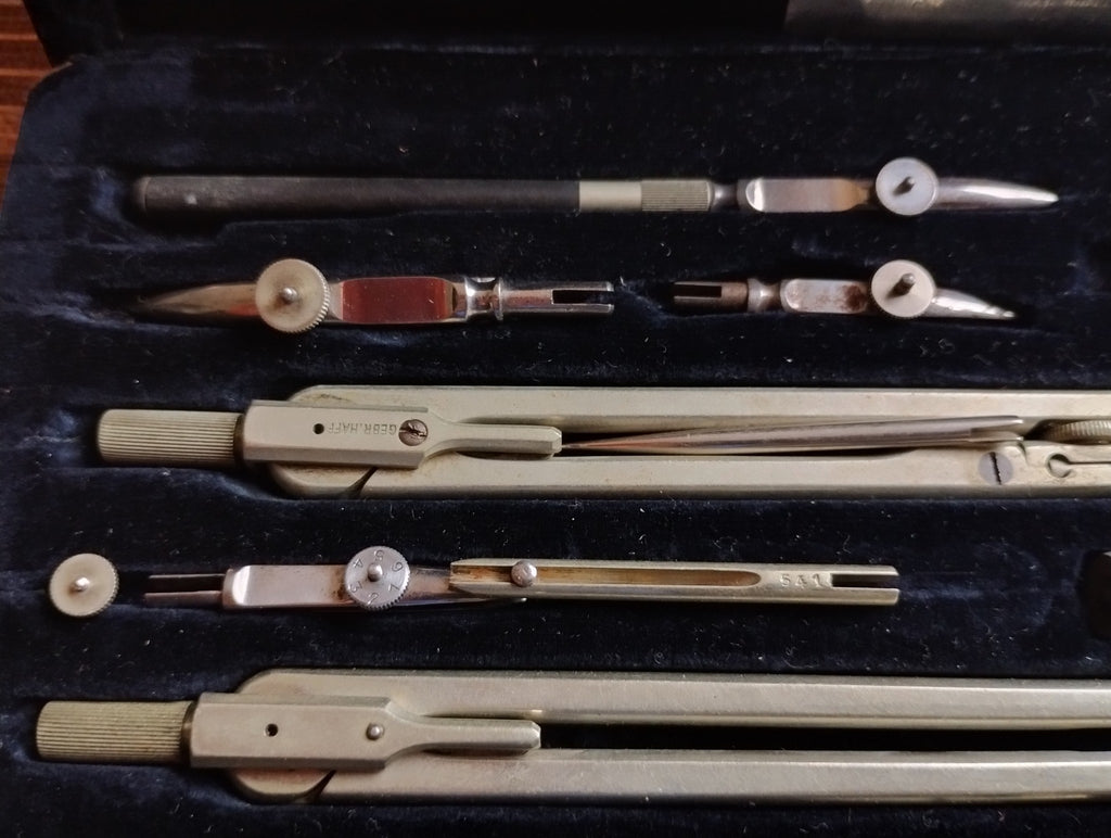 Close-up of metal compasses and drawing pen attachments in the dark fitted lining of the drafting set.
