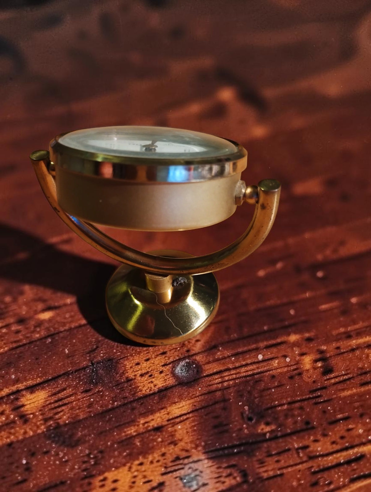 Vintage brass desk thermometer tilted backward in its curved support, showing the case and round base.
