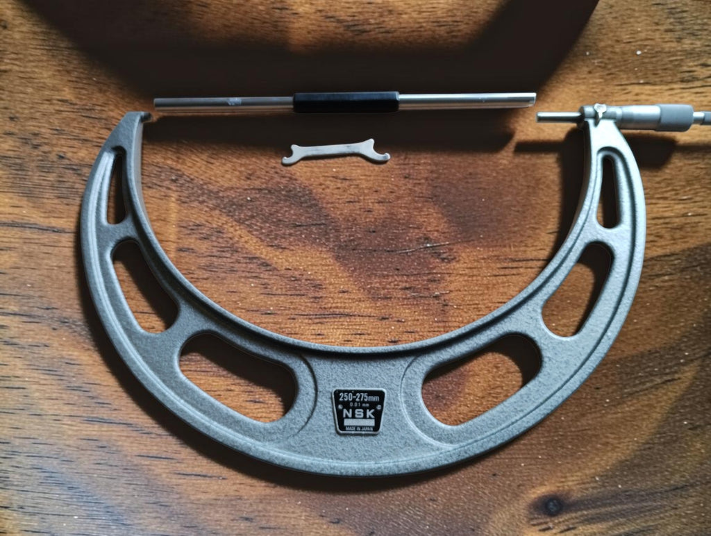 NSK 250–275 mm outside micrometer with gray hammertone frame, setting standard and adjustment wrench on a wooden surface.

