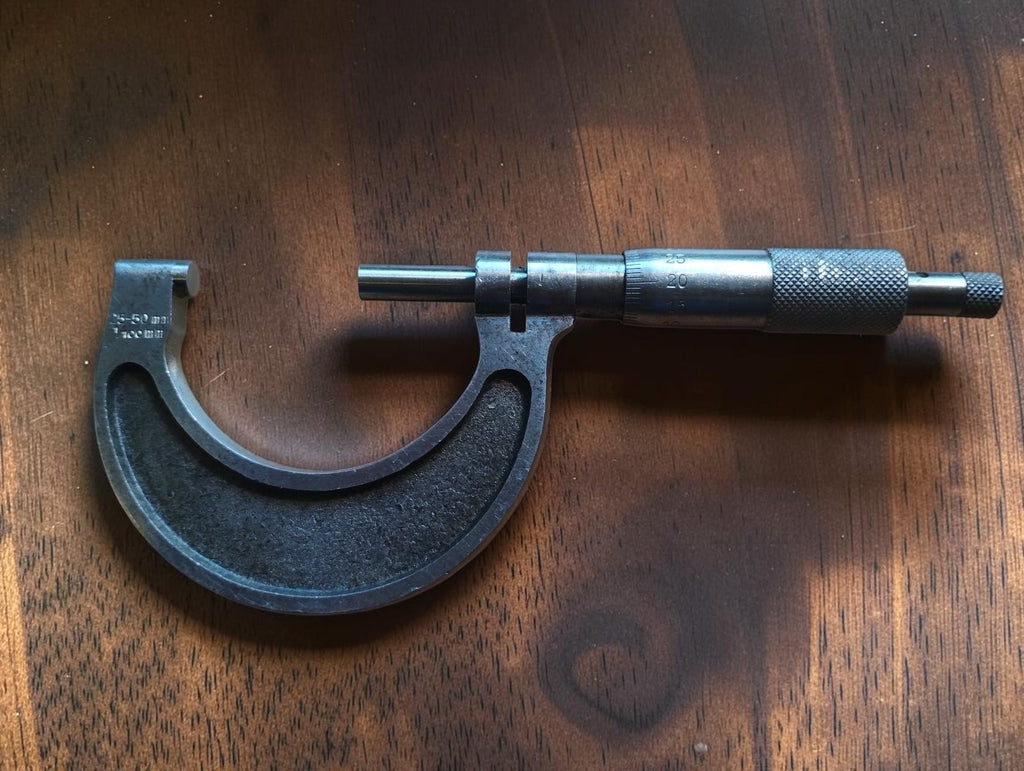 25–50 mm outside micrometer with a dark inset frame and knurled thimble, shown vertically on a wooden surface.
