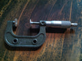 Vintage disc micrometer with a dark metal frame, round measuring faces and a graduated silver-tone thimble, shown vertically.
