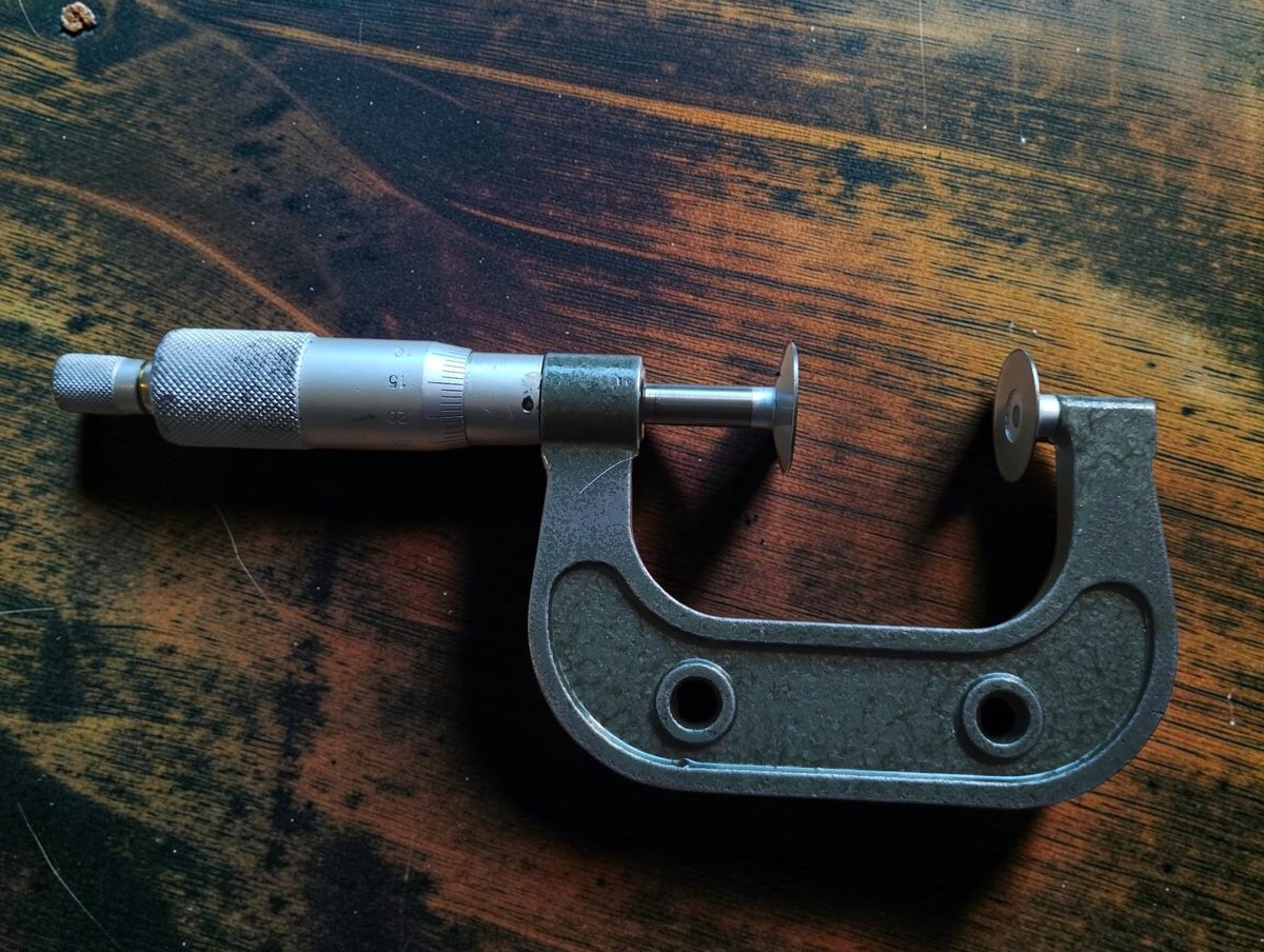 Full side view of a vintage disc micrometer showing its metal frame, two broad measuring discs and knurled adjustment thimble.
