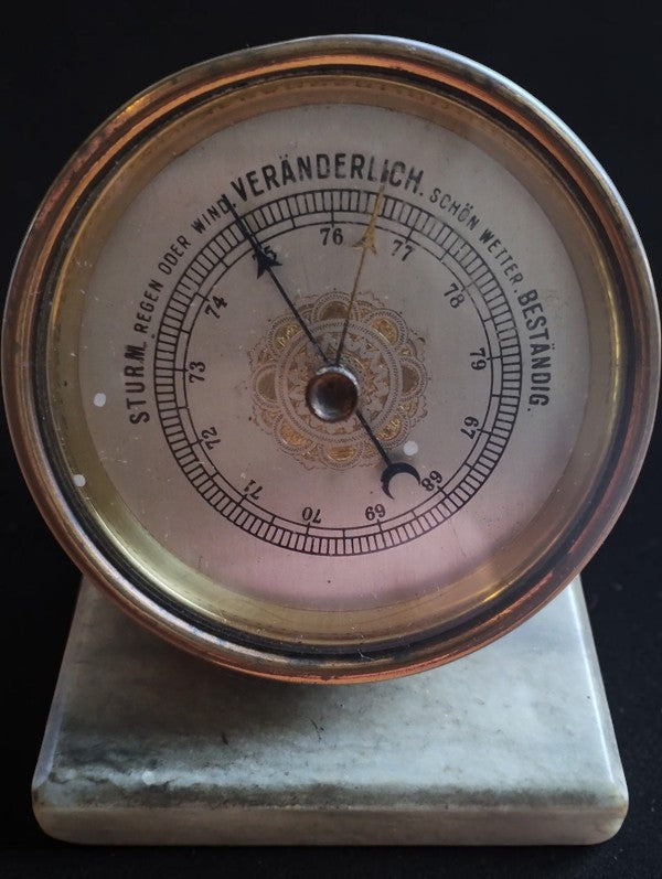 Front view of the brass aneroid barometer with decorative dial, visible age-related wear, and pale marble base.
