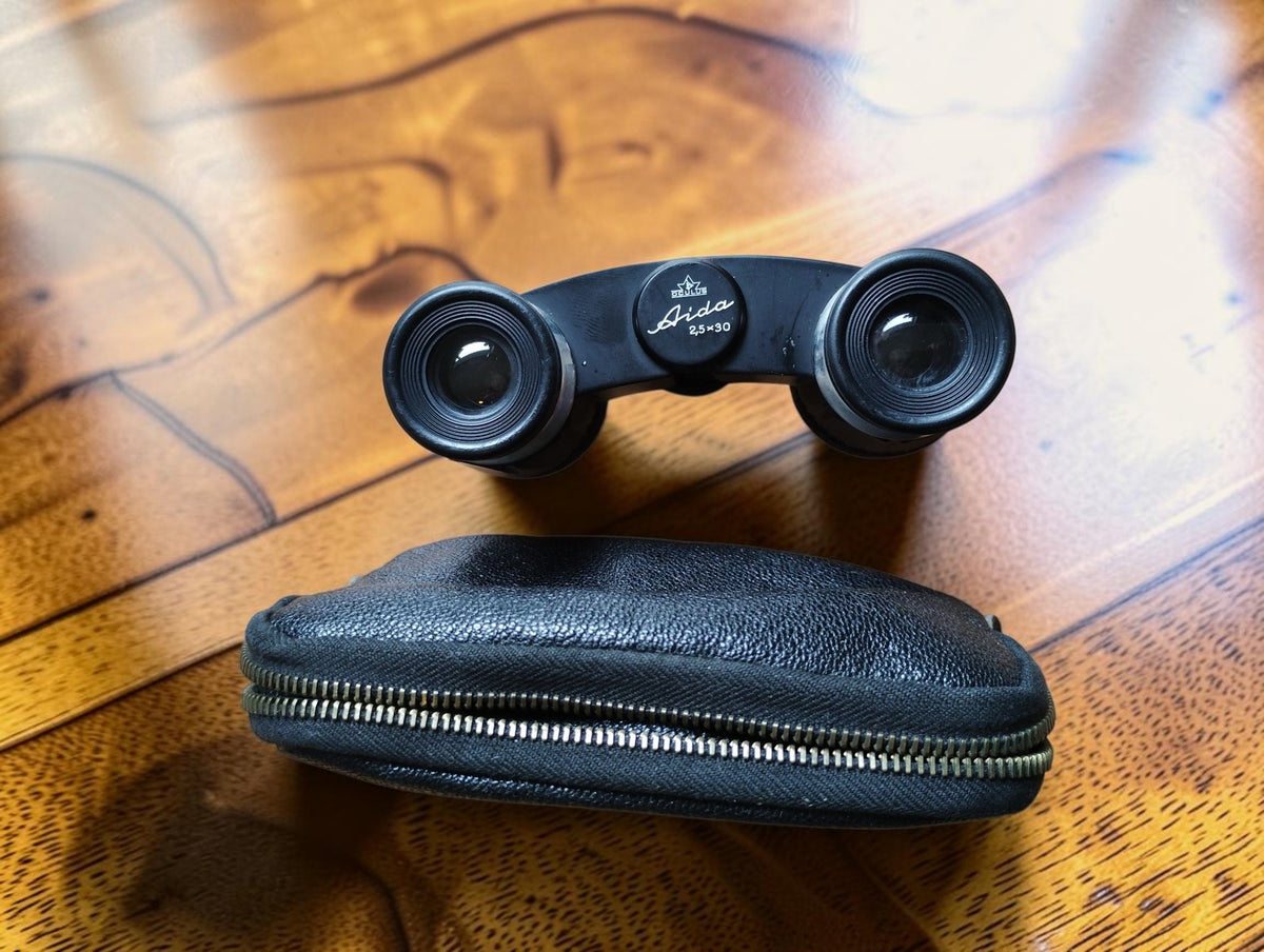 Vintage Aida 2.5 × 30 opera glasses with black zippered carrying case on a wooden table
