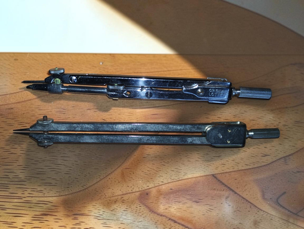 Two vintage metal drafting instruments with pointed tips and adjustment screws, shown outside their case
