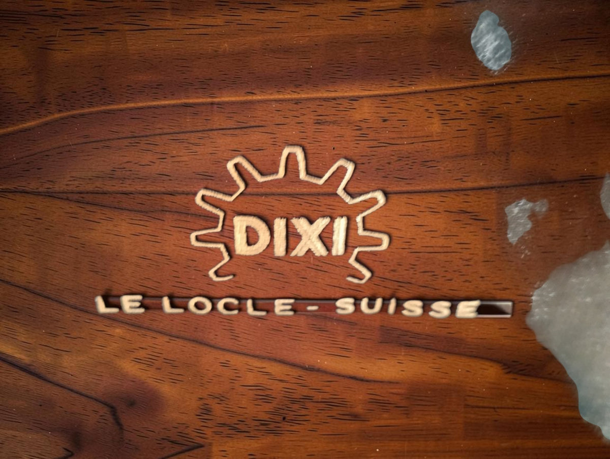 Close-up of the gold-colored DIXI gear logo and Le Locle Suisse lettering inside the watchmaking tool case lid

