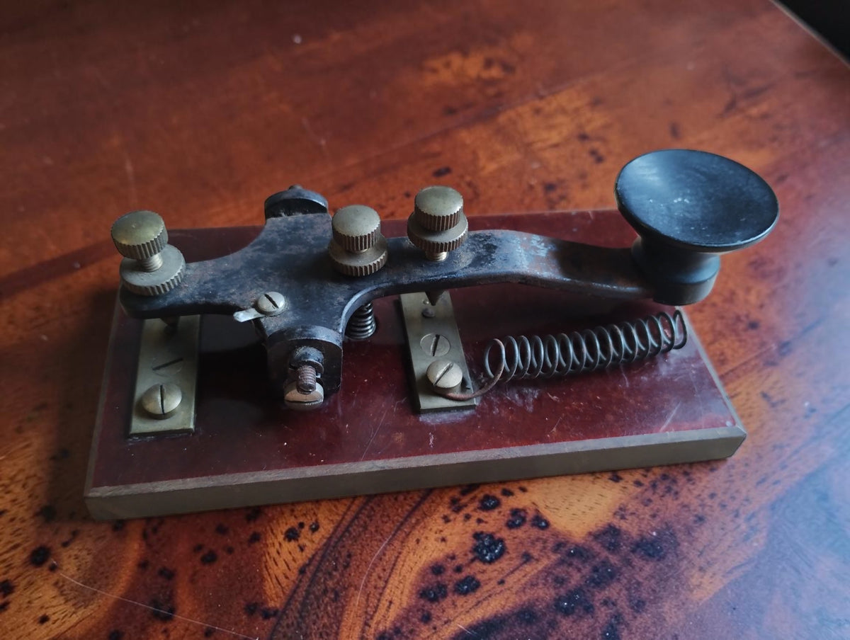 Antique Morse telegraph key on a reddish-brown base, showing the black operating knob, adjustment screws and long coil spring.
