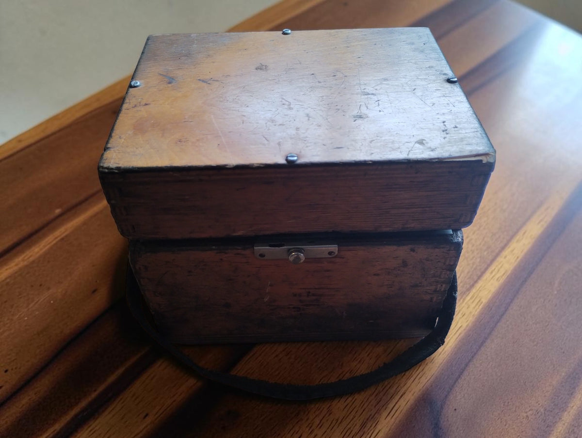 Closed wooden carrying case with aged patina, metal latch and black carrying strap for a vintage DC volt and amp meter.

