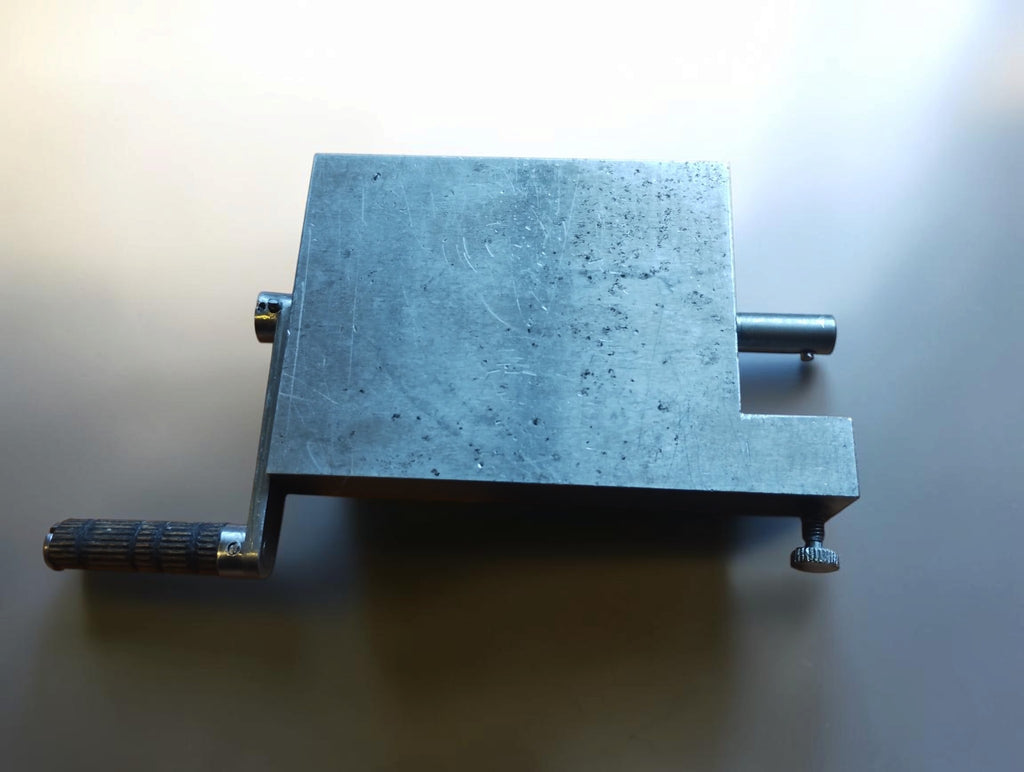 Top view of a metal hinge filing jig with a textured crank handle, projecting shaft and knurled screw.