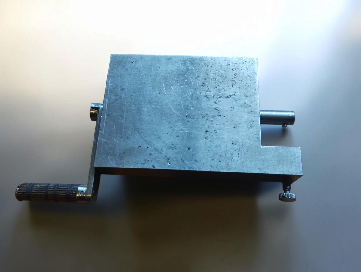 Top view of a metal hinge filing jig with a textured crank handle, projecting shaft and knurled screw.