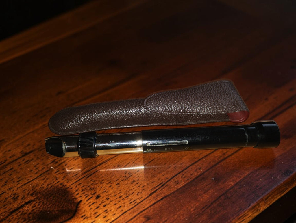 Zwarte vintage penmicroscoop met metalen details en zakclip naast het bruine etui
