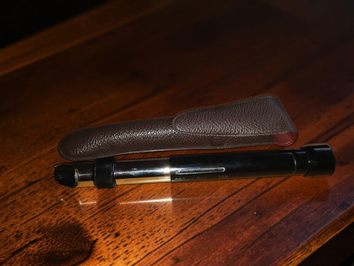 Zwarte vintage penmicroscoop met metalen details en zakclip naast het bruine etui
