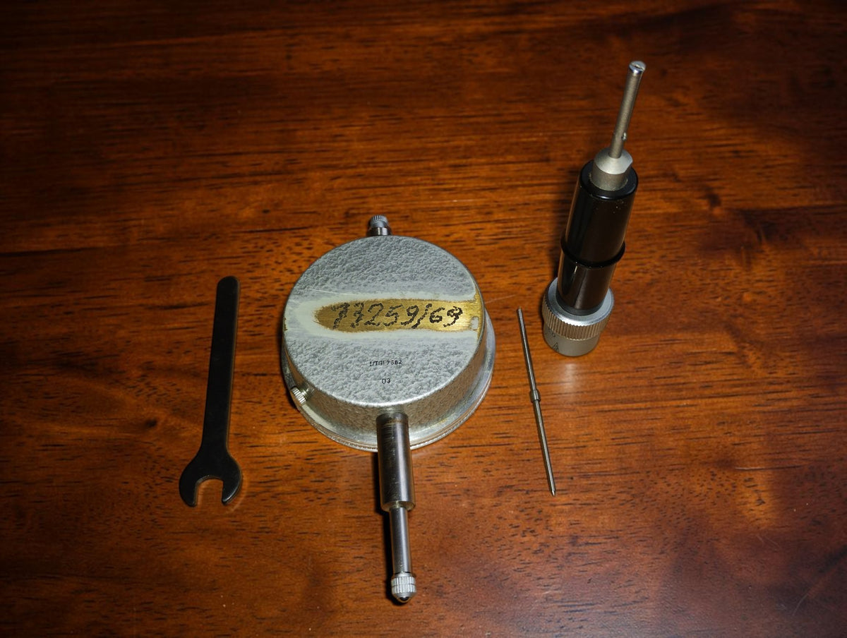Achterzijde van de INTO meetklok met handgeschreven markering op tape, naast de losse meetkop, sleutel en dunne pen.
