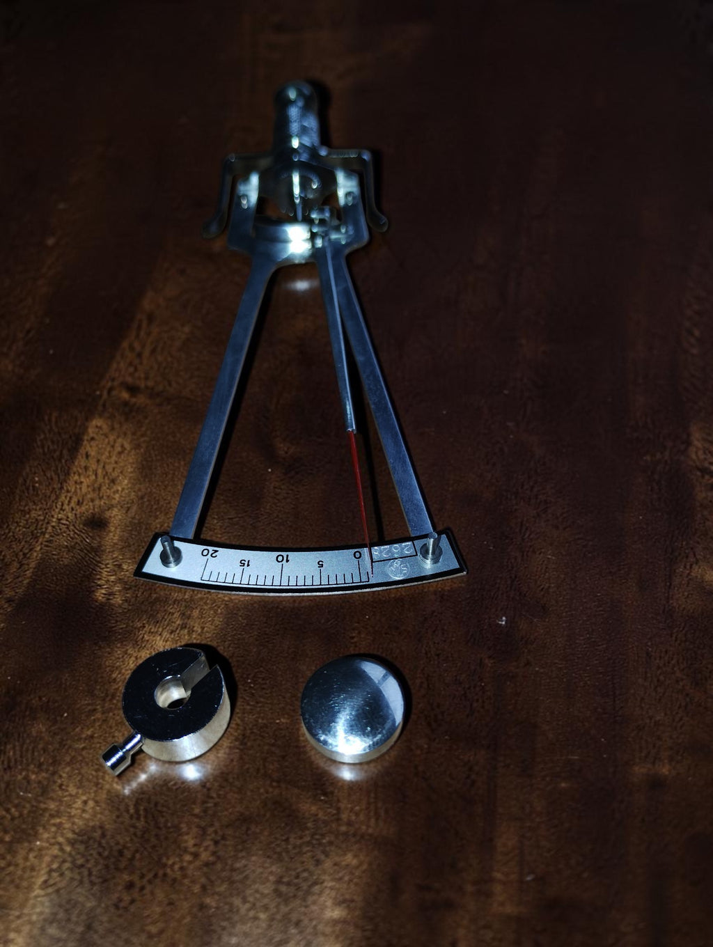 SK Schiötz tonometer on a wooden surface with two detached metal weights beside the numbered scale.
