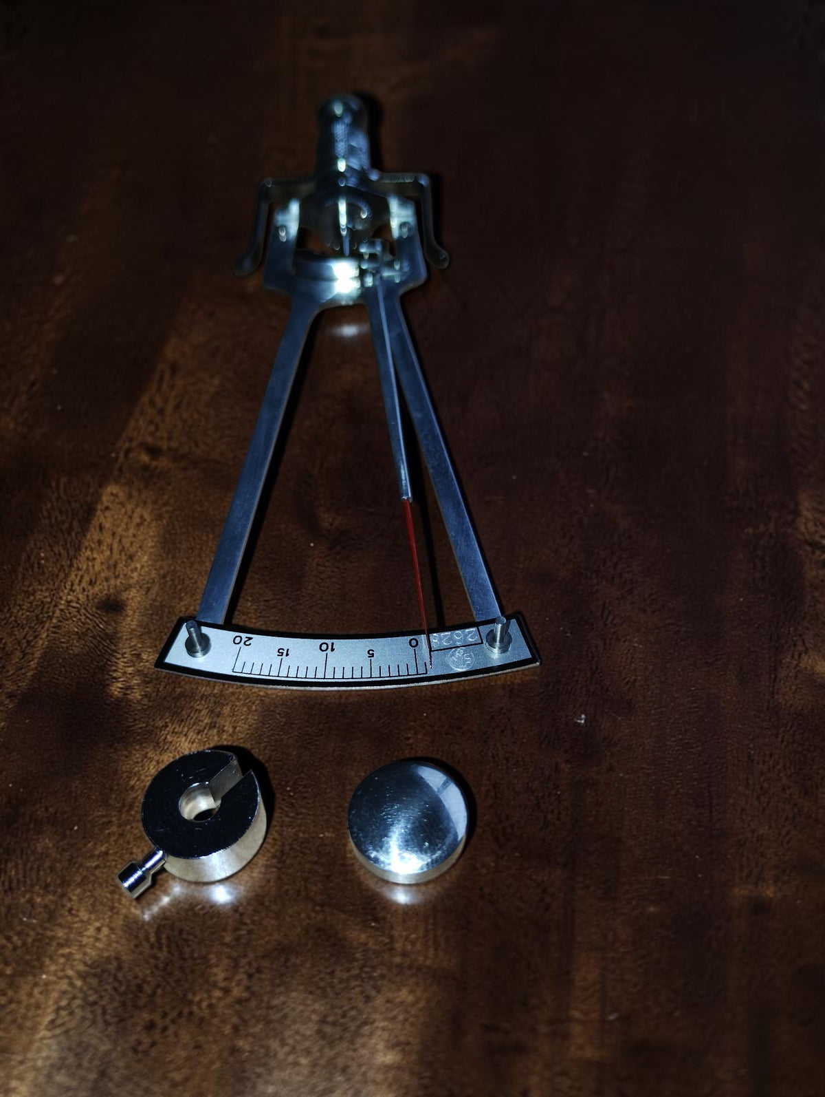 SK Schiötz tonometer on a wooden surface with two detached metal weights beside the numbered scale.
