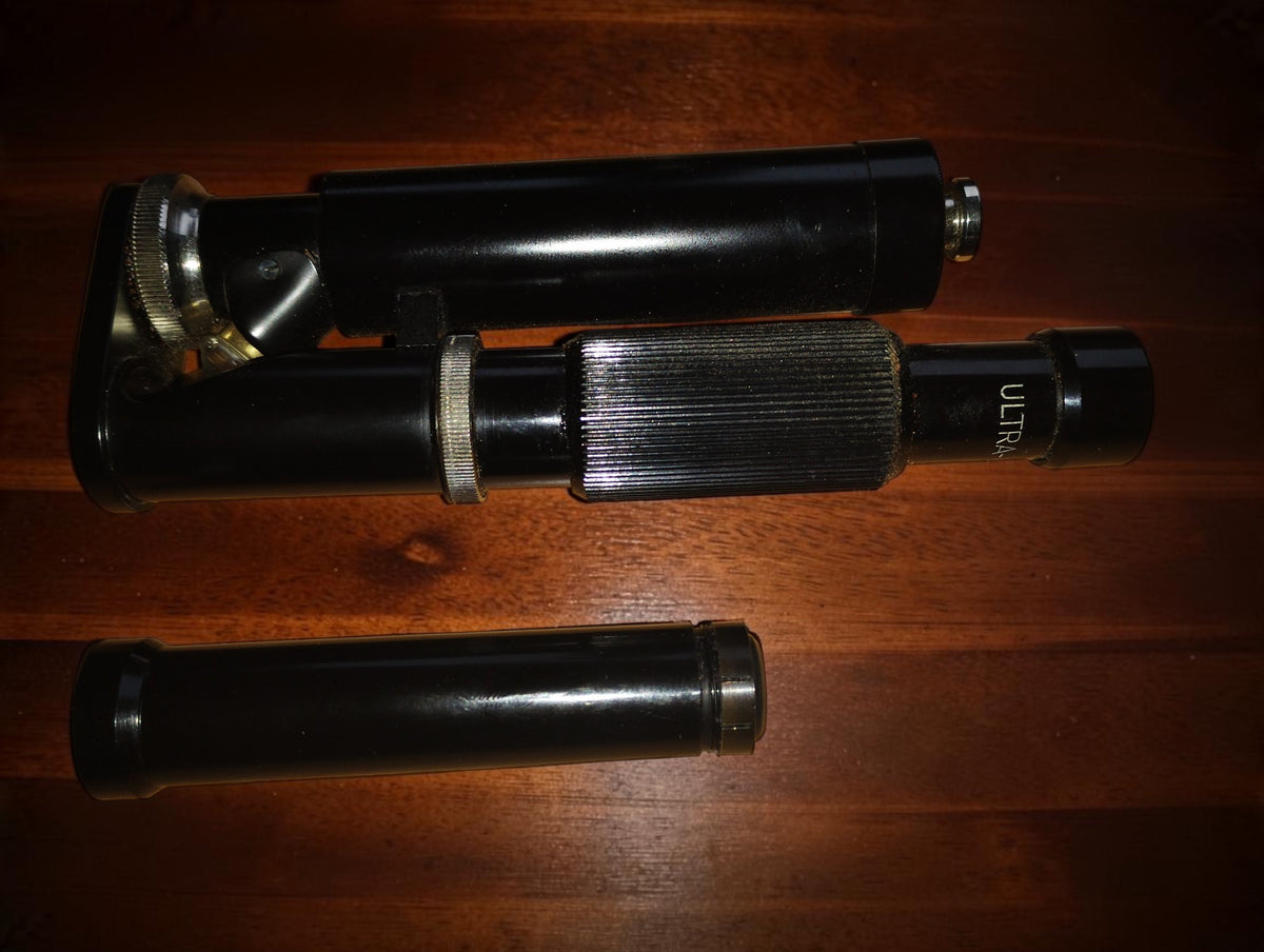 Ultra-Lomara pocket microscope lying horizontally on a wooden surface, with a separate black tube below.

