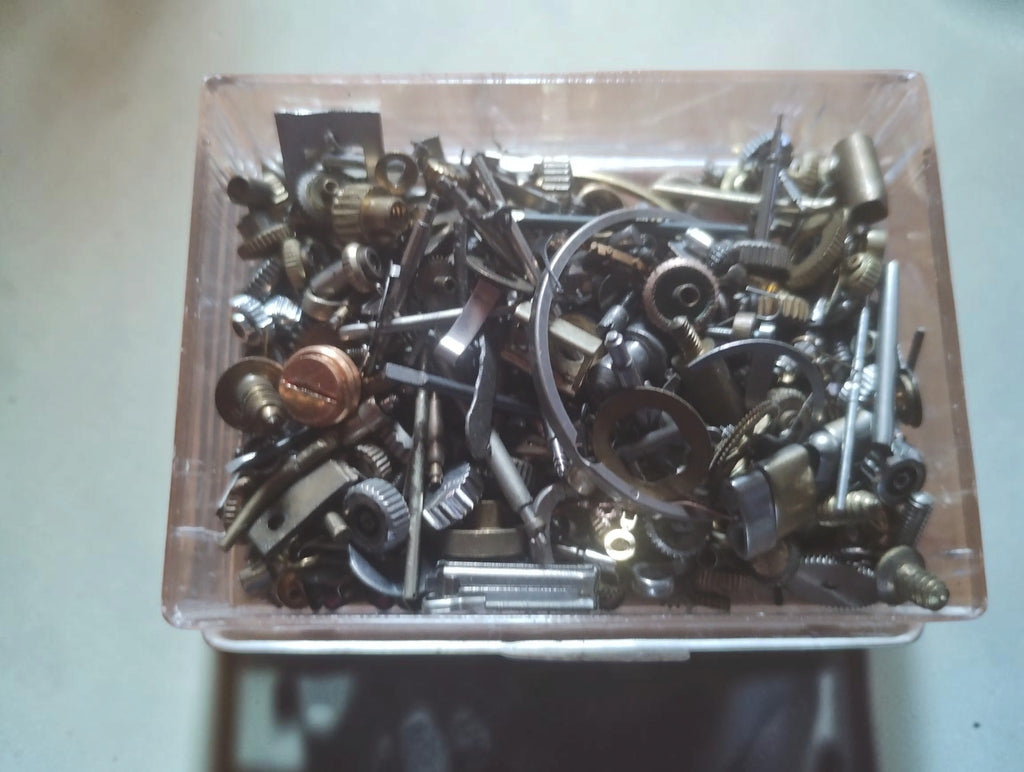 Close-up of assorted vintage watch movement parts, including small shafts, screws and toothed components in a clear box.
