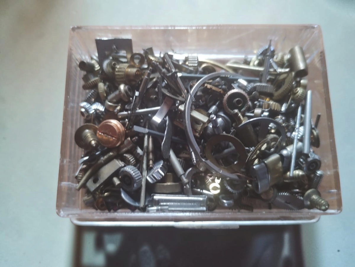 Close-up of assorted vintage watch movement parts, including small shafts, screws and toothed components in a clear box.

