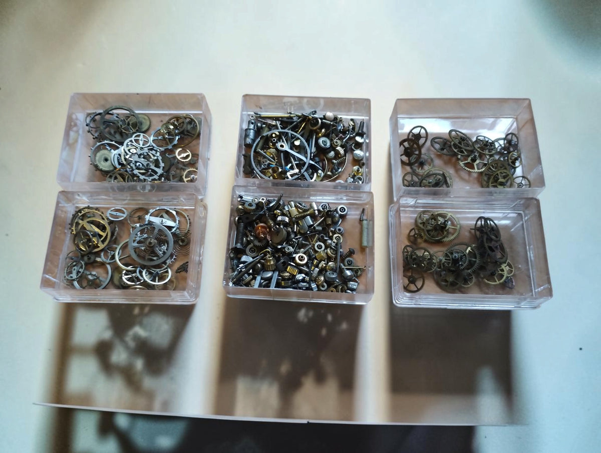 Overhead view of three open boxes and their lids holding vintage watch balance wheels, gears and small movement parts.

